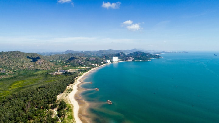 Aerial view of the long curving beach at Pak Nam Pran with forested hills, calm turquoise water and low-rise resorts along the shore