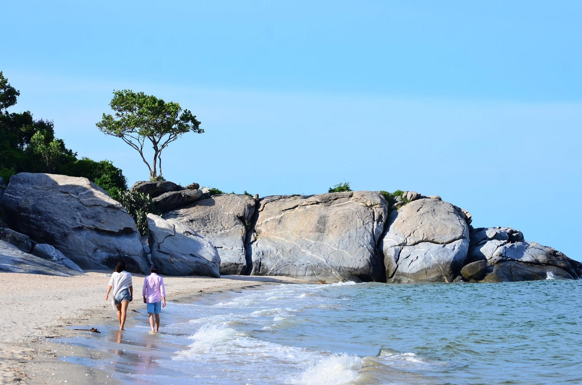 Two people walking along the shoreline at Sai Noi Beach beside large granite boulders