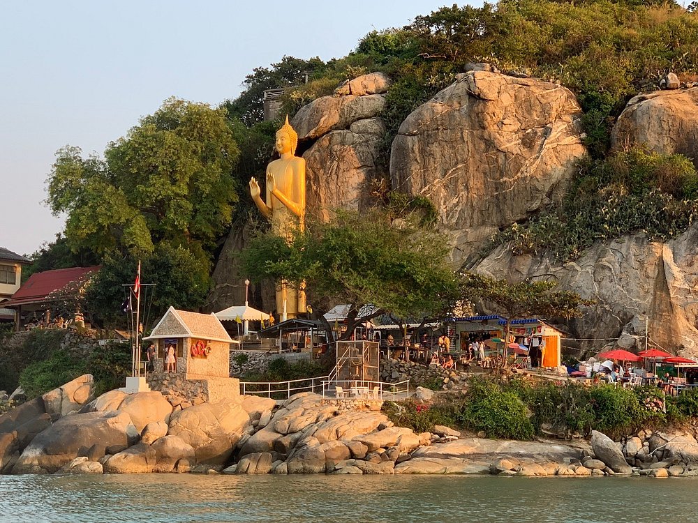 The golden standing Buddha at Wat Khao Takiab against a rocky cliff face above the water