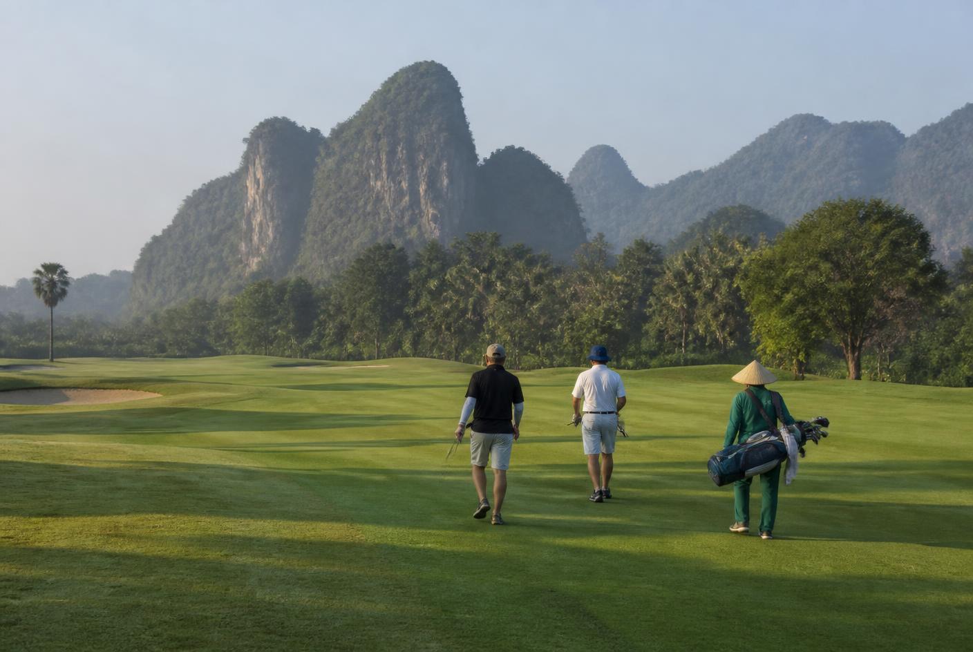 Two golfers and a caddie walking a fairway with limestone hills behind, early morning light